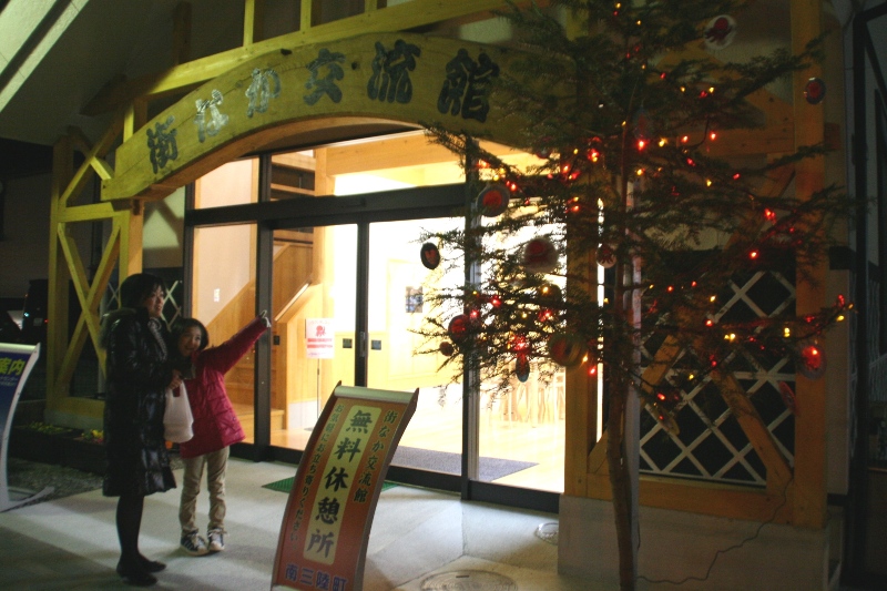 薄暗い街なか交流館の玄関横の木に赤い優しい光のイルミネーションが灯り、一組の親子が玄関前に立ち止まってイルミネーションを指さしている写真