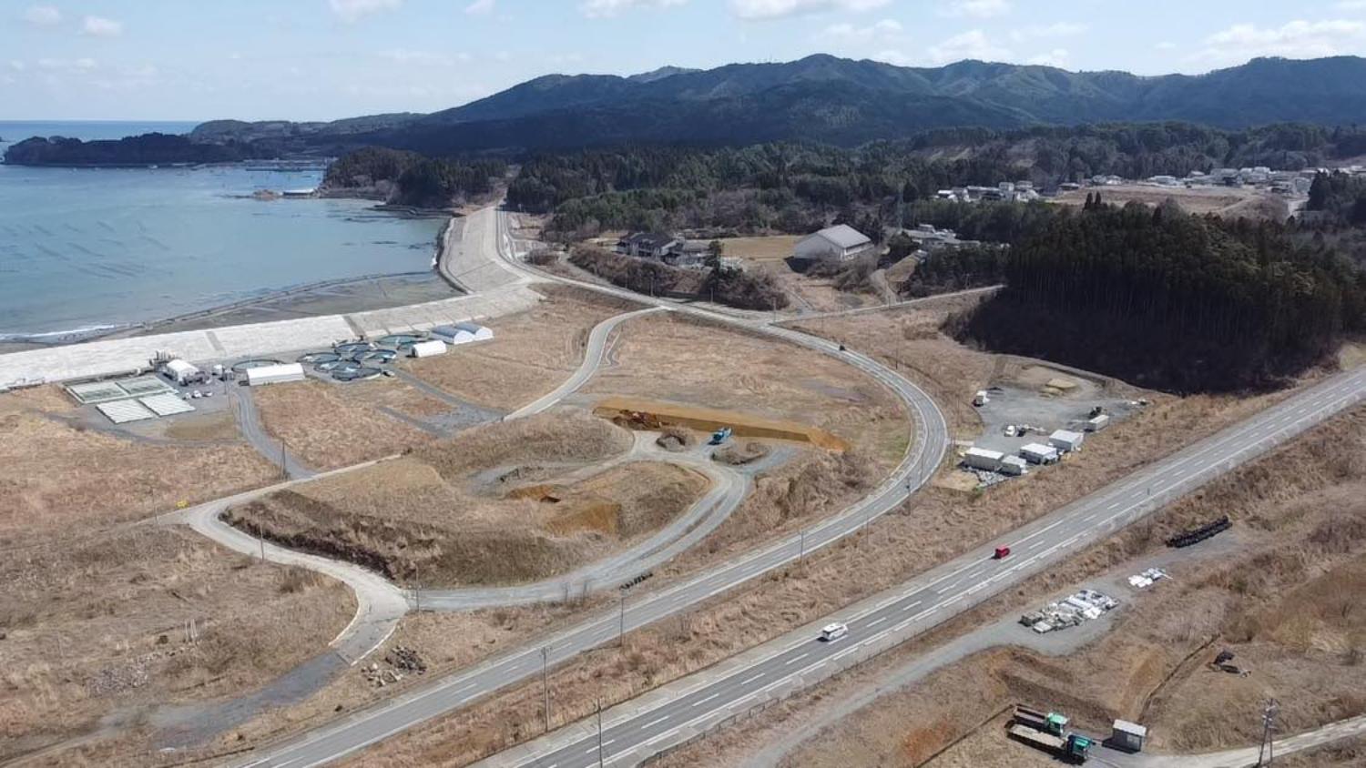 手前の道路と緩やかな丘、奥にはコンクリート製の大きな堤防と青い海、その周囲に小さな施設や建物が点在していて、復興のために整備された海岸とその背後の地域が見渡せる海沿いの昼間の風景を高台から定点カメラで撮影した写真