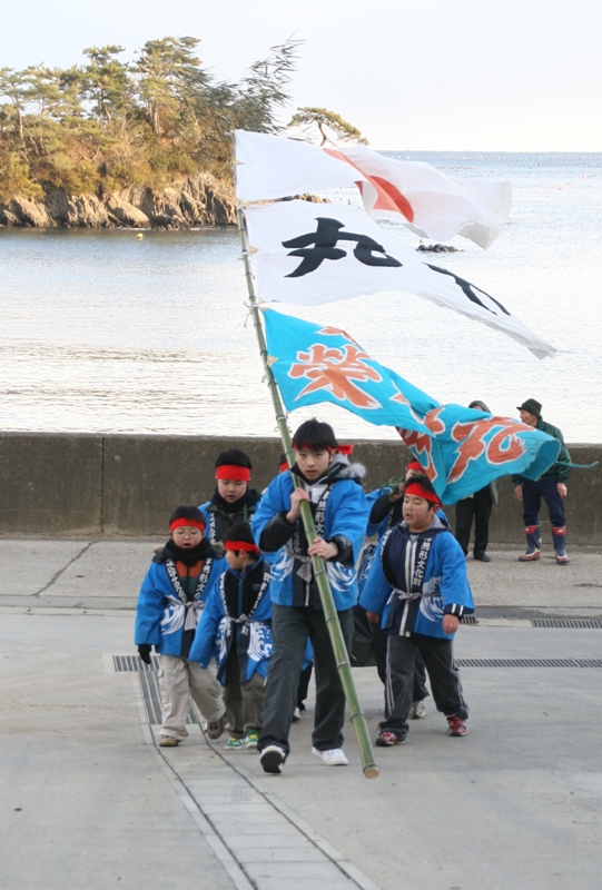 岸沿いの道を、青い法被に赤い鉢巻きを巻いた7人の子どもたちが歩いており、先頭の男の子は、3枚の旗が取り付けられた竹を担ぎ、そのあとに他の子どもたちが続いており、伝統行事「ささよ」に参加している様子を写した写真