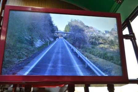 白い車線と両側にガードレールがあるまっすぐな道路が映し出され、道の両側には緑の木々が広がり、遠くには陸橋のような構造物が見えるモニター画面の写真