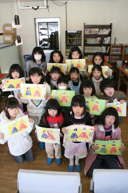 子どもたちそれぞれが作った色鮮やかなお内裏様とお雛様の貼り絵などの作品を両手で持ってカメラの方を向いて並んでいる集合写真