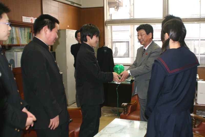 応接室で制服姿の志津川中学校の生徒会執行部の皆さんが訪れ代表の男子生徒が町長に緑色の募金箱のようなものを両手で手渡しており、生徒と町長が笑顔で向き合っている様子を捉えた写真