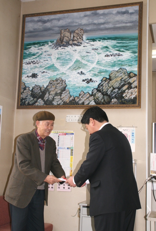 寄贈された「白波合掌」油絵の前で、受賞した佐々木久義さんと町長が向かい合って寄贈の受け渡しが行われている様子の写真