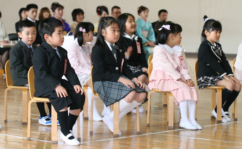 体育館に並べられた小さな椅子に真新しいスーツやワンピースを着た新一年生が緊張した面持ちで椅子に座っており、後方では保護者の方々が見守っている入学式の写真