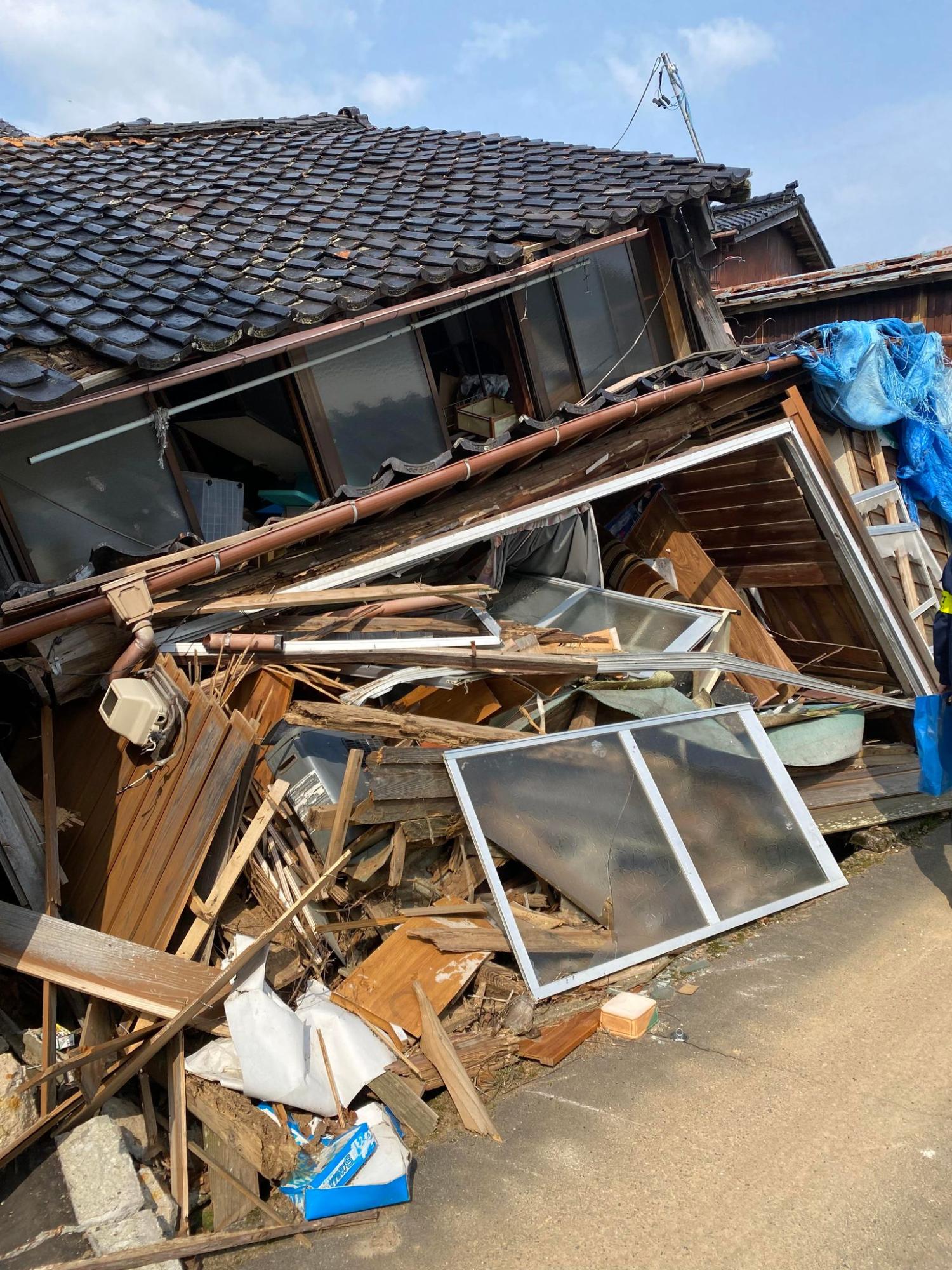 瓦屋根の家が大きく傾き、壁や窓、柱などが崩れて大量の木材やガラスが道路に散乱している写真
