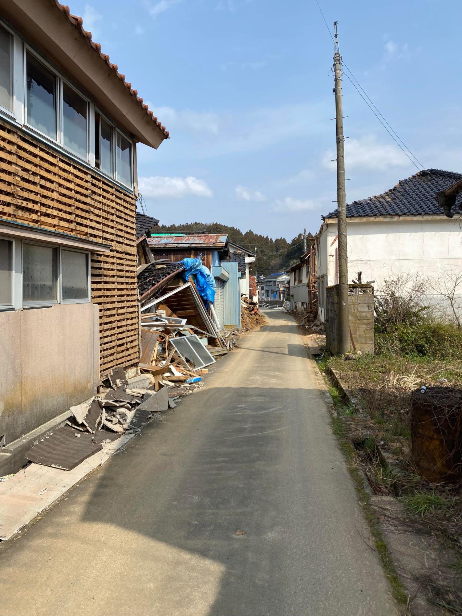 細い舗装された路地の両側に古い家屋が立ち並び、左側の建物の外壁や屋根が壊れて瓦礫や木材が建物の脇に積まれている様子の写真