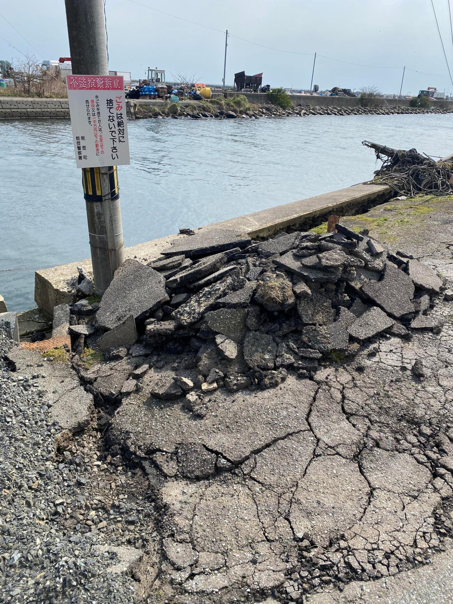 川沿いのひび割れたアスファルト道路に、壊れた舗装材が山のように積み上げられた写真