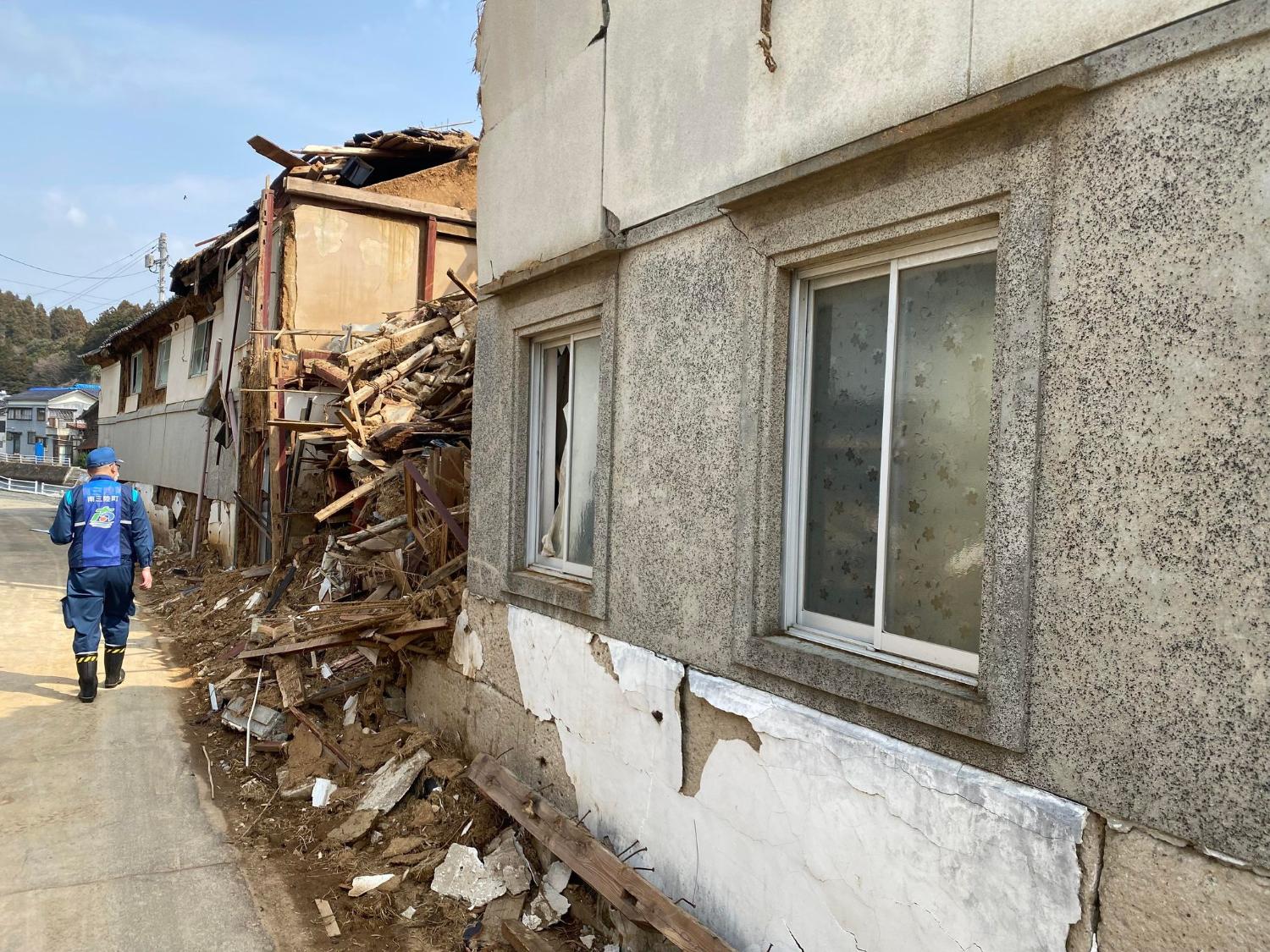 建物の壁はひび割れ、隣の家は木材や瓦礫がむき出しになっている場所を男性が観調査する様子の写真
