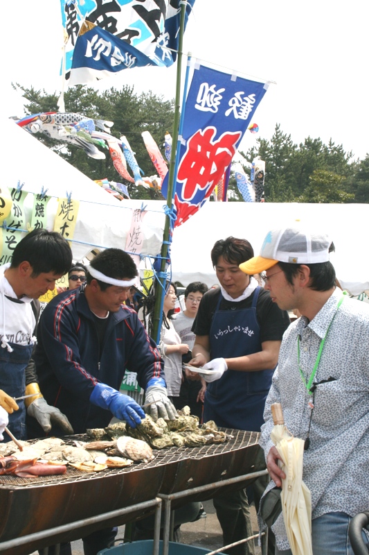 鯉のぼりが泳ぎのぼり旗が立つ海鮮物を扱う出店で、青い手袋をした男性が大きな鉄板の上で牡蠣やホタテなどの魚介類を焼いて調理しており、多くの来場者で賑わっている祭りの風景を捉えた写真