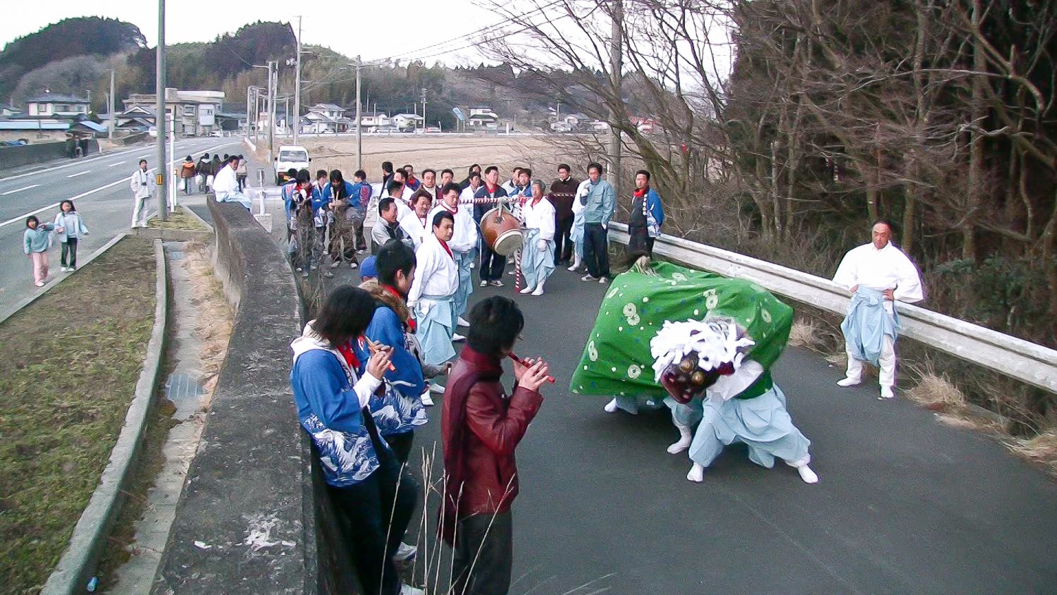 田舎の道路上で笛や太鼓を鳴らす大勢の人々に囲まれながら、緑の布に覆われた獅子舞が勇壮に踊っている様子の写真