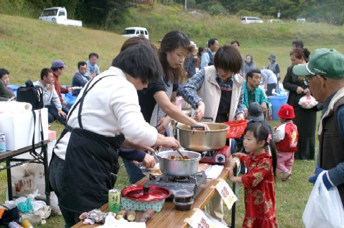 広々とした草地の野外で複数の大人が料理を準備し、テーブルの周りに集まって食事や交流を楽しんでいるイベント風景の写真