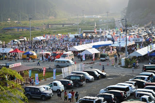 夕暮れ時の広い駐車場と河川敷に設けられた会場に白いテントが並び、たくさんの人々が屋台やステージを楽しんでおり、奥には川と橋、手前にはびっしりと並んだ車が見える、イベント会場の全体のにぎわいを遠景からとらえた写真