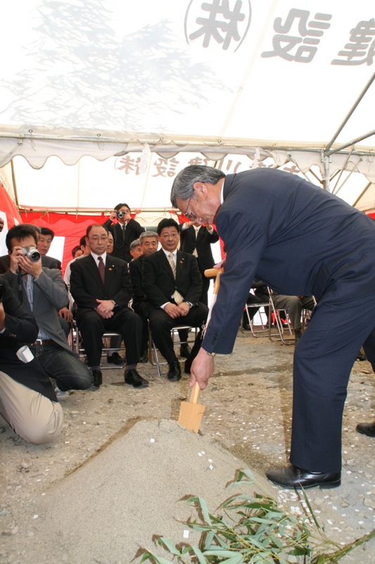 紅白幕がまかれた白いテントの下で参列者が見守る中、スーツ姿の関係者の男性が木製の鍬を使って砂山に手を入れる起工の儀式を行っている戸倉小学校体育館起工式の写真