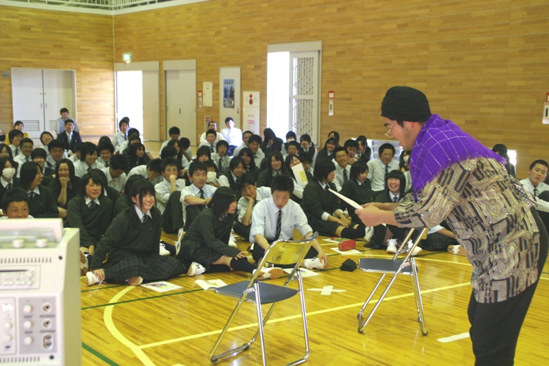 制服姿の多数の高校生が座って聴講する中、紫色のスカーフを巻いた男性が寸劇を披露している認知症サポーター養成講座の様子の写真