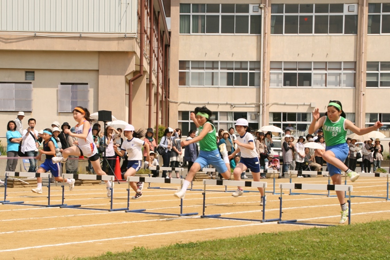 学校のグラウンドで観客に見守られながらハードル走の競技に挑んでいる小学生体育大会の写真