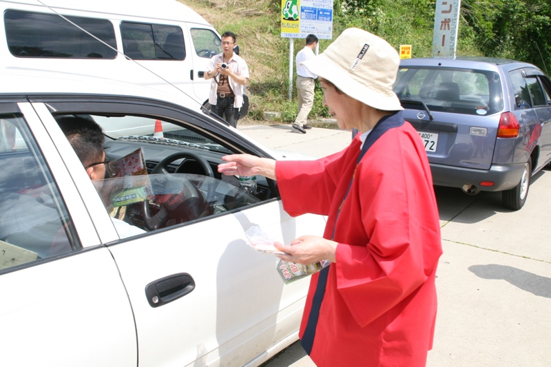 赤い法被と白い帽子をかぶった高齢の女性が、停車した白い乗用車の運転席に安全運転を呼びかけるチラシを手渡している交通安全啓発活動の写真