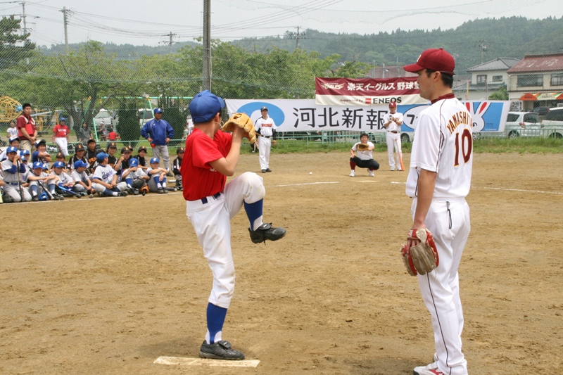 数十名の児童が見守る中、代表の児童がピッチング練習で投球フォームをとっており、楽天ジュニアコーチが側で指導している野球塾の様子の写真