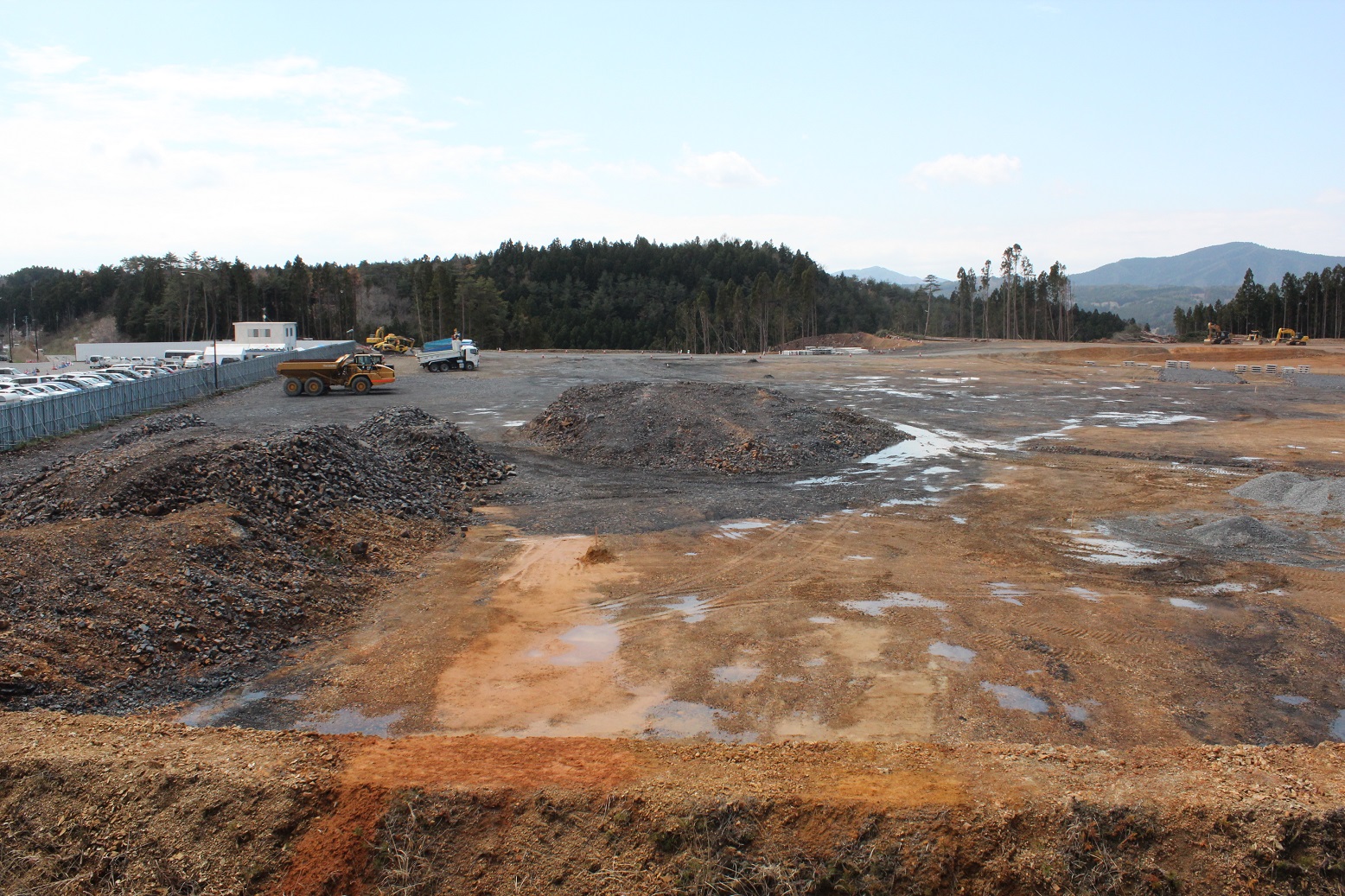 雨上がりの造成地に水たまりが点在し、中央には砕石の山とダンプカーやショベルカーが見え、左手には仮設建物と駐車車両が並ぶ写真