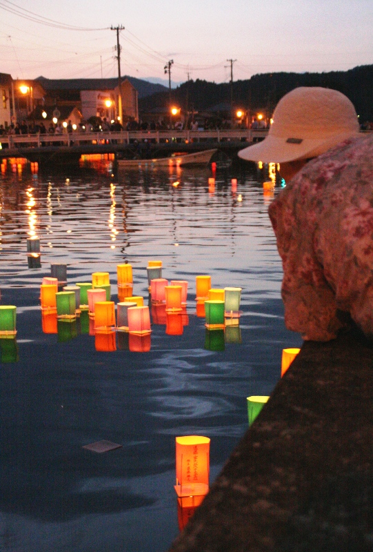 夕暮れの八幡川で、オレンジ・緑・ピンクなど色とりどりの灯ろうが水面を流れ、川沿いの建物の灯りと遠くの山並みが背景に映り、欄干から灯ろうを見守る人の姿も見える流灯会のの様子の写真