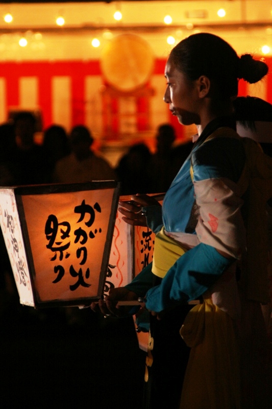 浴衣を着た女性が『かがり火祭り』と書かれた灯りのともった提灯を手に持ち、背景には祭りの装飾や灯りが見える夜の祭りの様子を写した写真
