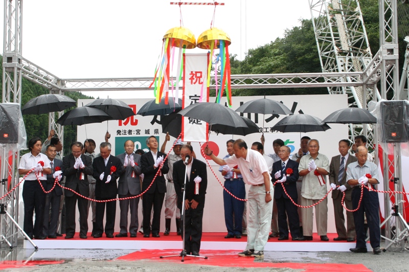 赤い絨毯が敷かれた屋外の式典会場で、くす玉が割られて「祝 定礎」の幕が垂れ下がる中、スーツ姿の多数の関係者が黒い傘を差して並び、マイクの前で一人の男性が挨拶のため頭を下げている、払川ダム定礎式の様子の写真