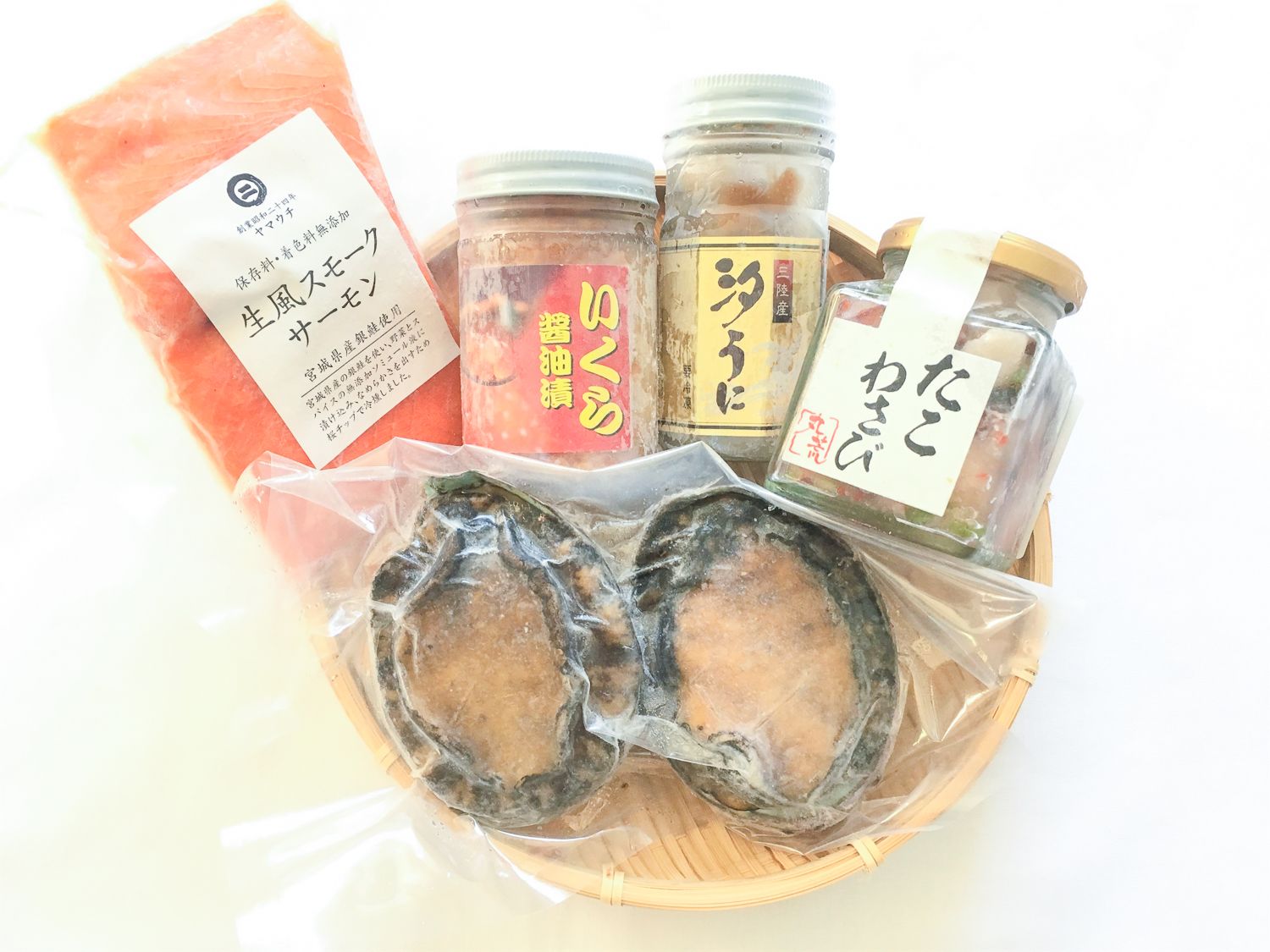 スモークサーモンやいくらの醤油漬け、汐うに、たこわさびなどの海産加工品とアワビが竹ざるに美しく盛られている南三陸町のふるさと納税返礼品セットの写真