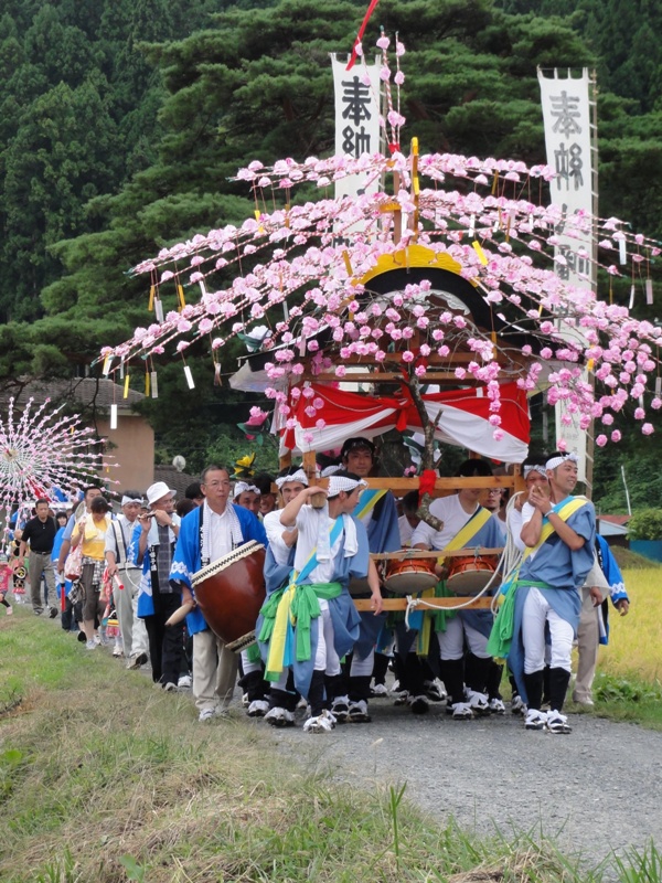 田んぼに囲まれたあぜ道で、華やかな牡丹の花飾りを施された華やかな山車を複数人が担ぎ、太鼓や笛などの楽器を演奏する人々とともに進む、入谷打囃子の祭りの様子の写真