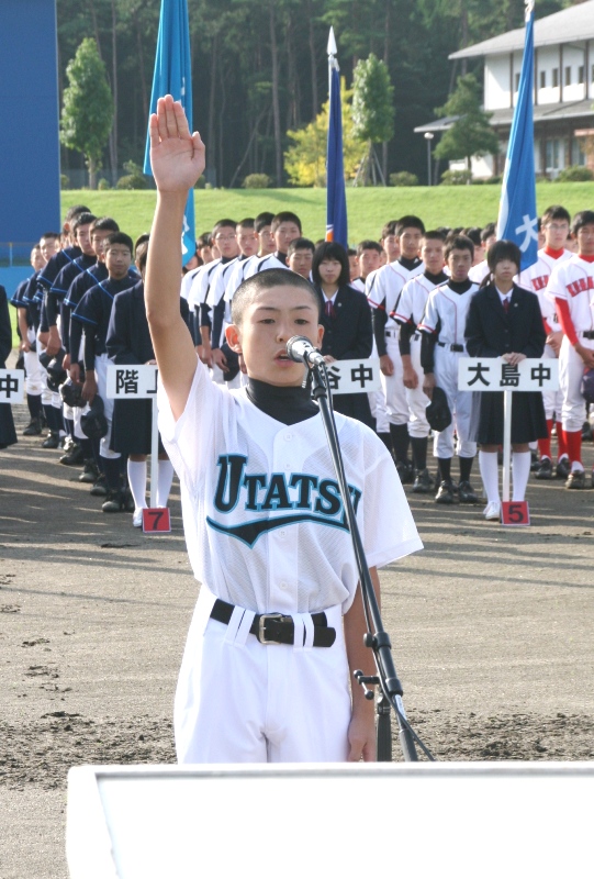 各校の旗やプラカードを持った選手たちが整列している屋外の会場で、歌津中学校野球部キャプテンの浅野祐介さんが右手を真っ直ぐに上げてマイクの前に立ち、選手宣誓を行っている様子の写真