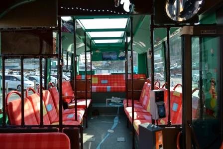 赤いチェック柄の座席が並び、大きな窓から自然光が差し込む車内で、横向きと後ろ向きの座席が混在した停車中のバスの内部を写した写真