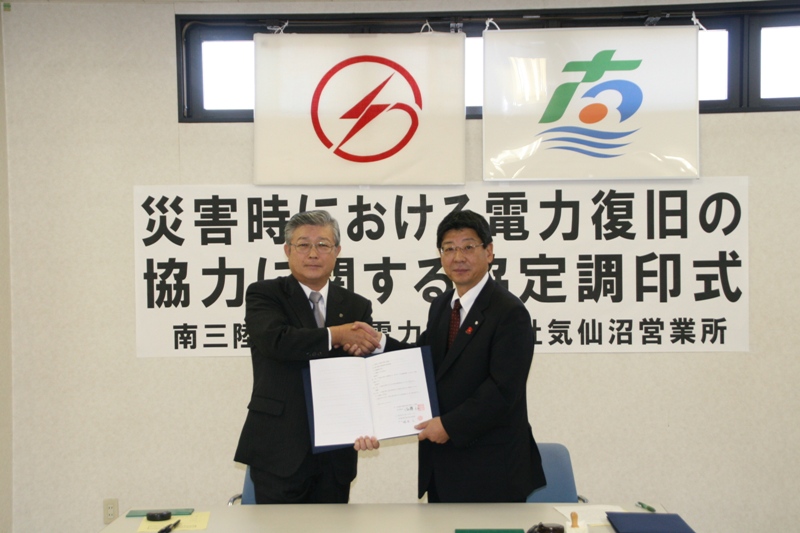 「災害時における電力復旧の協力に関する協定調印式」の幕が掲げられる中。町長と東北電力会社の代表者が協定書を一緒に持ち、固い握手を交わしている調印式の様子の写真