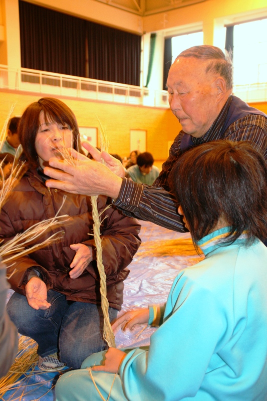 藁を束ねた縄を高齢の男性が両手で捩じ上げ、参加者の親子がその手元を真剣な表情で見つめている親子しめ縄づくり体験の様子の写真