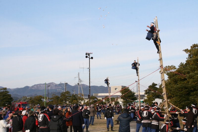 屋外の会場で山々を背景に、複他の団員が縄で梯子を支えている三本の高い梯子の上から消防署員が餅を空中に投げ、下に集まった人々がそれを受け取ろうとしている餅まきのの様子を写した写真