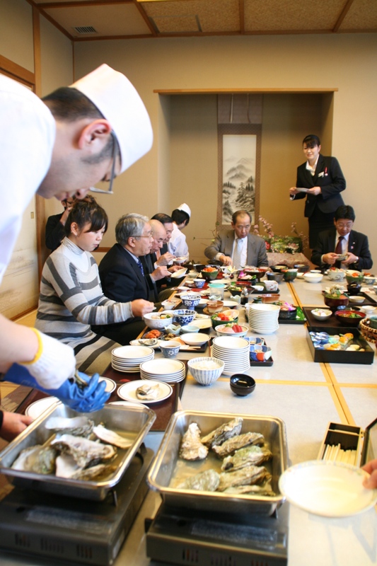 和室の畳の上に低いテーブルが設置された会場で、調理師が手前で牡蠣などの食材の準備をする中、海産物などの料理が机の上に並び、町長や関係者の方々が試食を楽しんでいる様子の写真