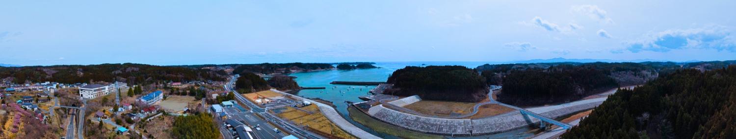 湾の入口には防波堤が見え、左側に住宅地、右側に山林と堤防が広がっている、海に面した小さな湾とその周辺の集落や山々を一望できる昼間の風景を高台から定点カメラで撮影した写真