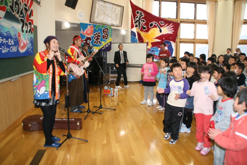 法被姿のマウンテンマウスのお二人が室内の会場でギターを演奏しながら歌い、子どもたちが笑顔で手を叩いたりしながら一緒にコンサートを楽しんでいる様子を写した写真