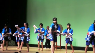 青と白の市松模様の揃いの法被を身にまとった子どもたちがの舞台上で手ぬぐいのような道具を手にして踊りを披露している戸倉浜甚句（とぐらはまじんく）の郷土芸能発表の写真。