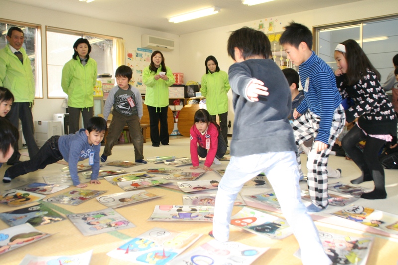 黄緑色のお揃いの上着を着た補導員協会の方々が笑顔で見守る中、中央の女性が札を読み上げており、室内に並べられた大きなかるたの絵札の中から読み上げられた絵札に向かって子ども達が駆け寄ってきているかるた大会の様子の写真