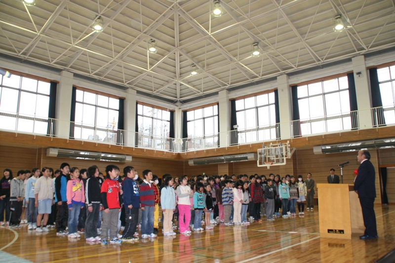 真新し体育館に全校児童の皆さんが整列しており、演台の前に立つ赤い花飾りをつけたスーツ姿の男性が挨拶をしている様子の写真