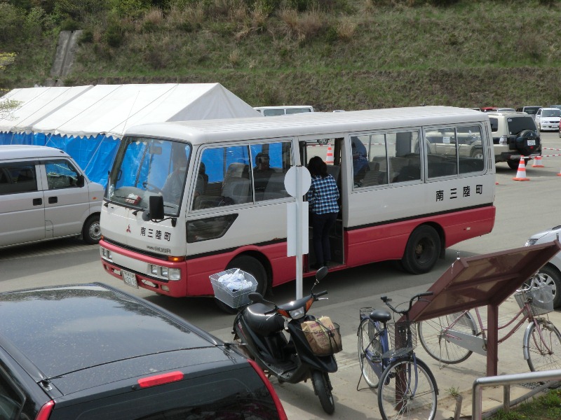 南三陸町と書かれた紅白のツートンカラーのマイクロバスが屋外のバス停前に停車している写真