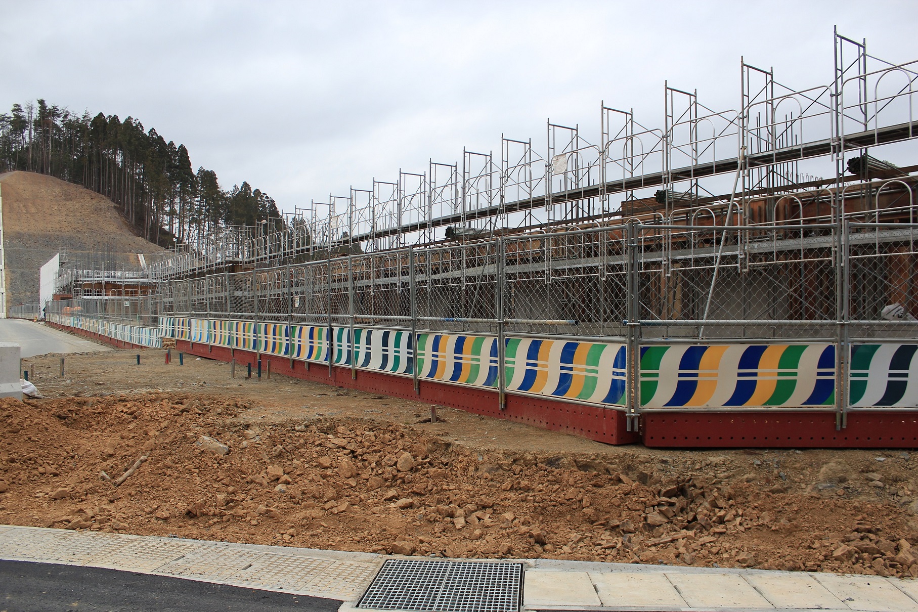 鉄筋が組まれた構造物の上に足場が設置され、手前にはカラフルな模様が描かれた仮囲いと掘り起こされた赤土が広がる、山あいの建設現場の様子を捉えた写真