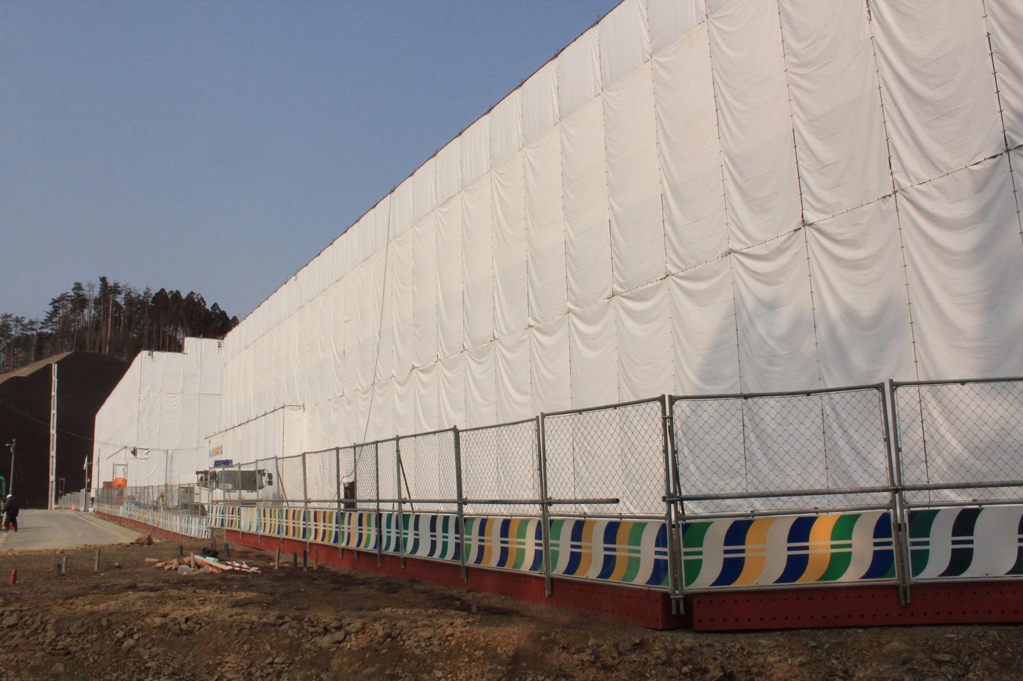 白いシートで全面が覆われた大規模な建設現場の外壁沿いに、色とりどりの模様が描かれたバリケードとフェンスが設置され、左奥には木々が立ち並ぶ斜面が見える写真