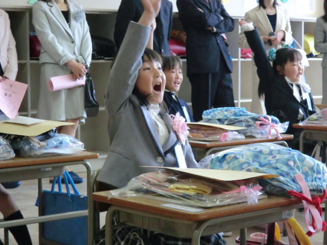 教室でスーツ姿の保護者が見守る中、新一年生が席についており、嬉しそうな笑顔で元気よく右手を挙げて返事をしている入学式の教室での風景を写した写真