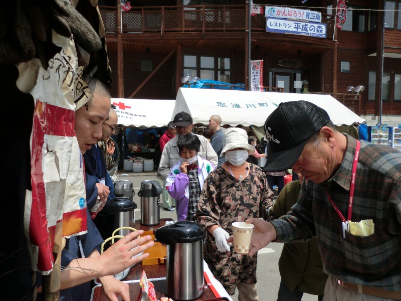 テーブルにポットが並んでおり、青年僧有志の皆さんが高齢の男性に紙コップに入ったお茶を手渡ししている様子を写した行茶ボランティアの写真