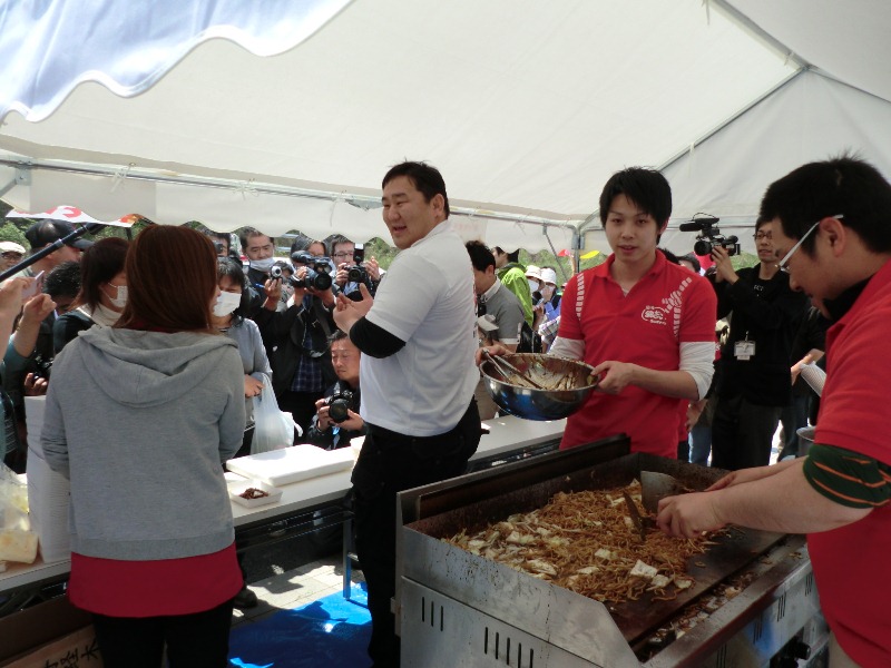 報道陣と多くの一般客に囲まれたイベント会場のテントの下で、後方の鉄板ではスタッフが焼きぞばを作っており、白いTシャツ姿で笑顔を見せる元横綱の朝青龍さんが写っている写真