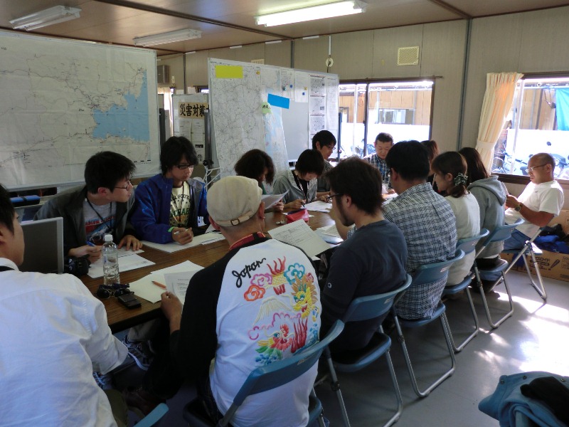 大きな地図が貼られたホワイトボードがある室内に長机が設置され、10数名の方が長机を囲んで各自資料に真剣に目を通しながら打ち合わせをしている最終打ち合わせの様子の写真