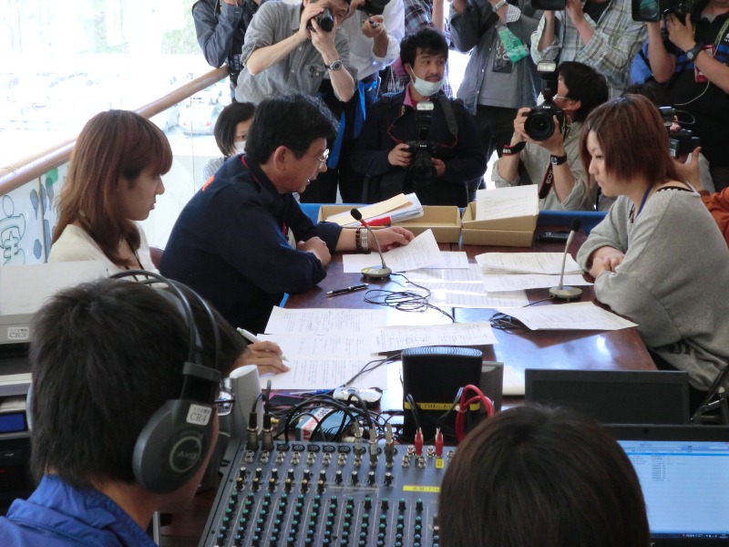 多数の報道関係者がカメラやビデオカメラを構えて取り囲む中、南三陸町長が原稿を手にマイクを前に話し、両隣の女性が聴き入っている南三陸災害エフエムのラジオ放送の様子を写した写真