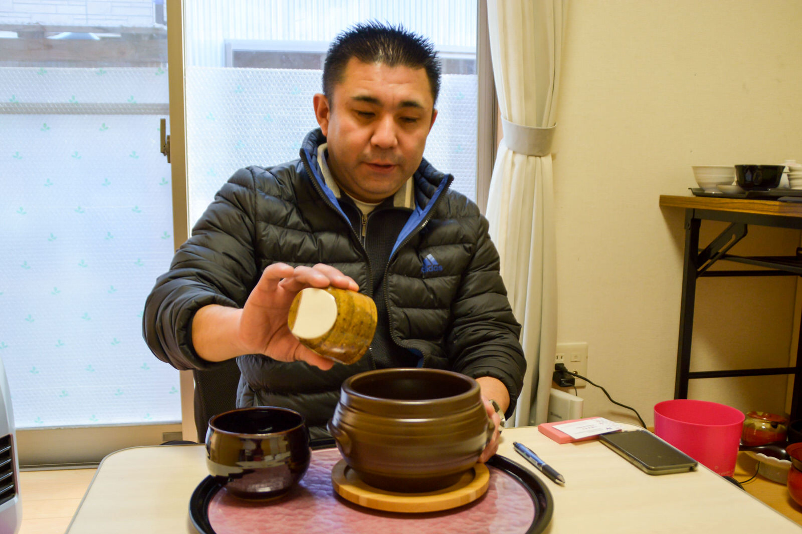 室内のテーブルにタコつぼを模した大小の器が乗ったお盆が置かれており、黒いジャンパーを着た後藤さんが大きい方の器に小皿を傾けて何かを入れている様子の写真