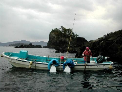 曇天の下、海に浮かぶ小型漁船に3人の人が乗り込み、海面に向かって体をかがめるなどして漁作業を行っているアワビ漁業の様子の写真