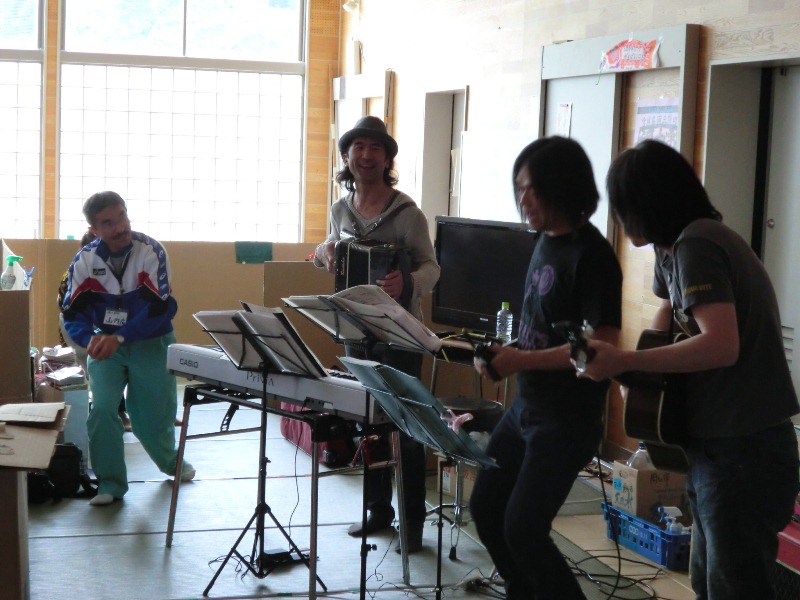 避難所の会場で、ソウルフラワーユニオンのメンバーがキーボードやアコーディオン、ギターなどの楽器を持って楽しそうに演奏している様子の写真