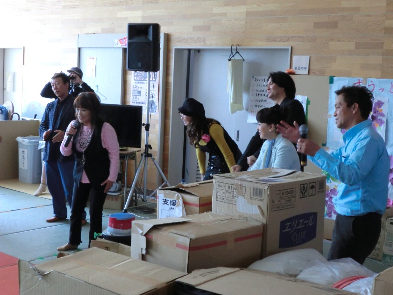 段ボール箱が積まれている避難所の会場で、金沢明子さんがマイクを持って話をしており、周りには橋幸夫さん、山本リンダさん、ジョン健ヌッツォさん、ジュディ・オングさん、清水あきらさんなども一緒に話を聞いている様子の写真